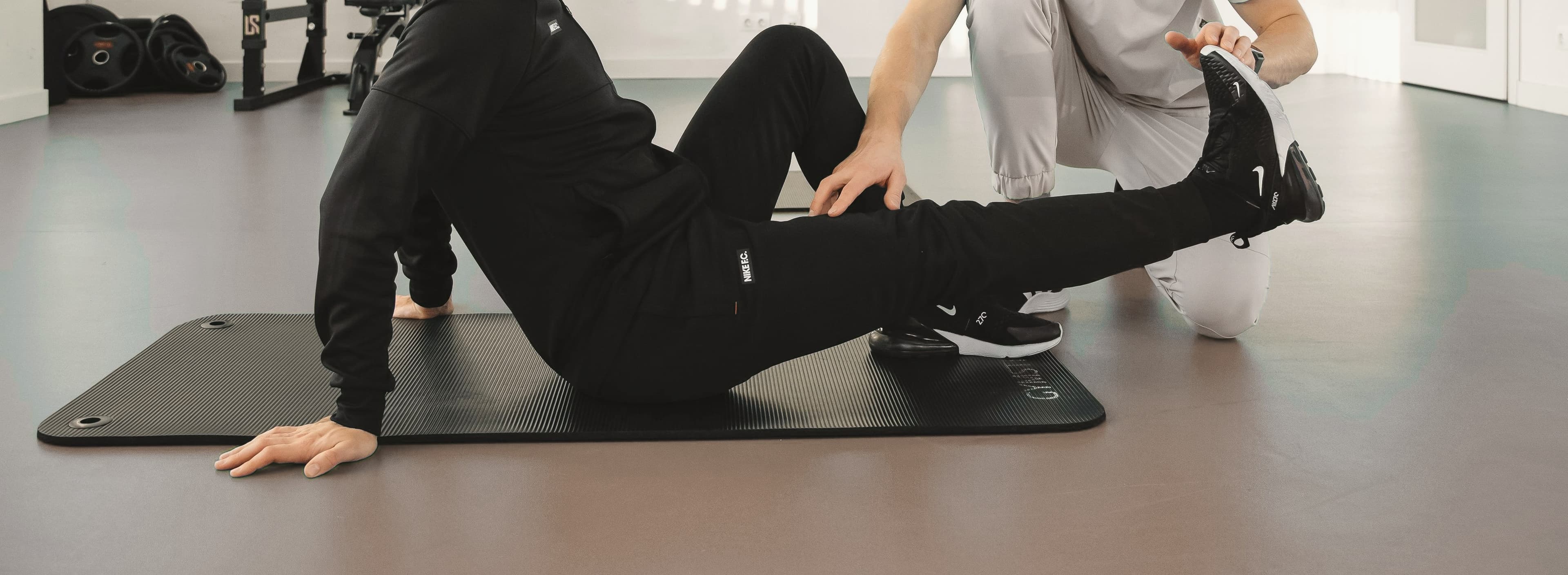 A physical therapist's hands guiding a client's raised leg during a stretch, on a mat in a bright therapy studio