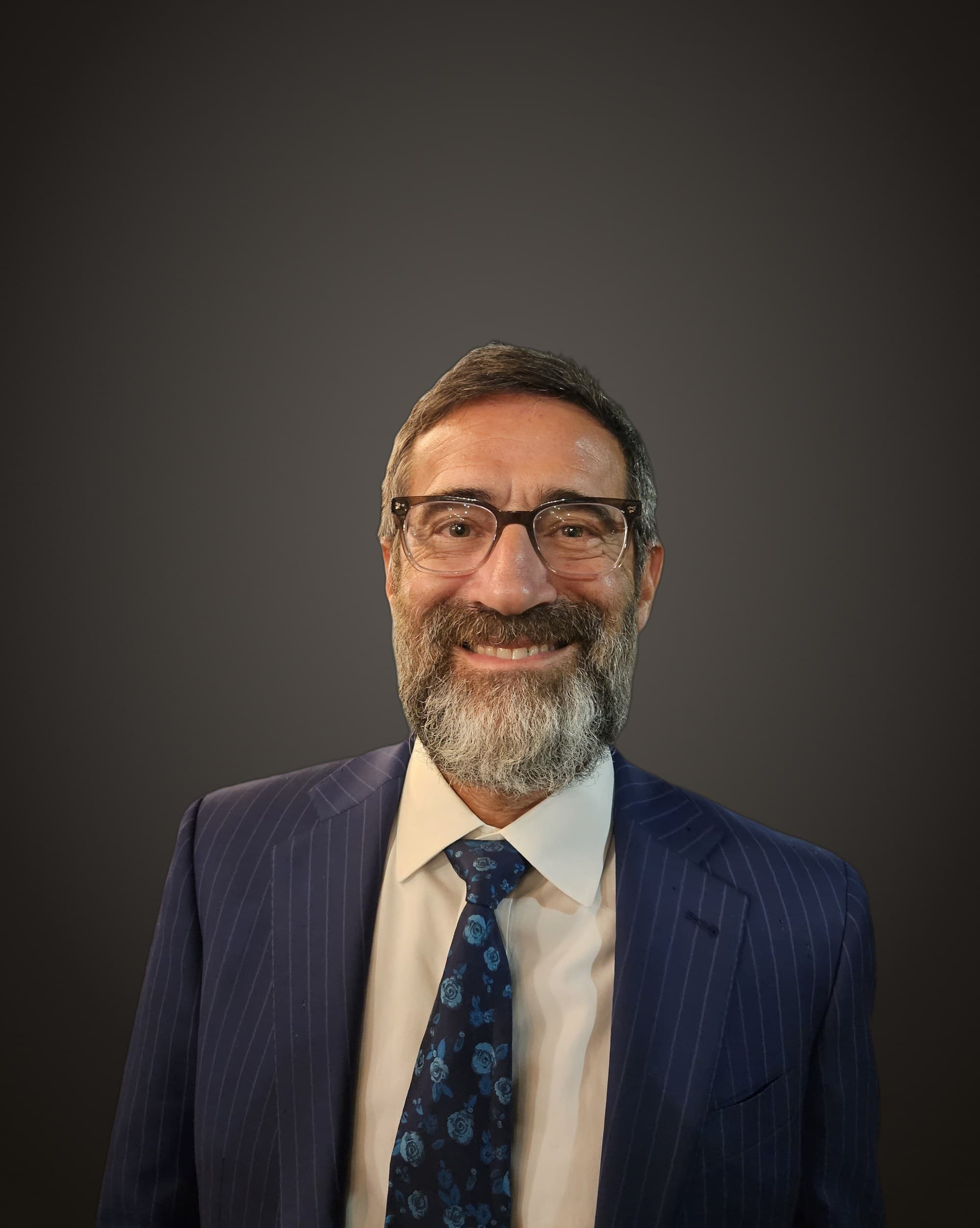 Chaim Backman, PT, smiling in a navy pinstripe suit and glasses against a deep charcoal studio ground