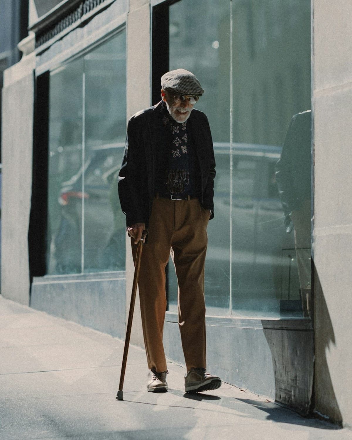 An elderly man in a flat cap using a cane, pausing on a New York sidewalk